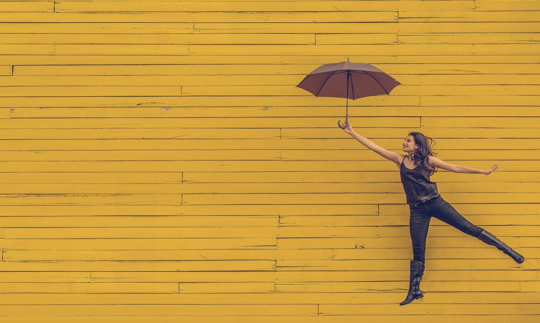 Vrouw springt in de lucht met een bruine paraplu tegen een achtergrond van gele planken. (Foto: Pexels | Pixabay.com)